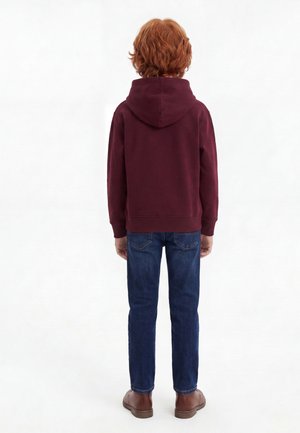Enfant aux cheveux roux, debout de dos, portant un sweat à capuche bordeaux, un jean bleu et des chaussures marron sur fond blanc uni.