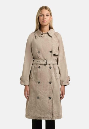 Kvinne iført en beige dobbelspent trenchcoat med belte, stående rett opp mot en ensfarget hvit bakgrunn.