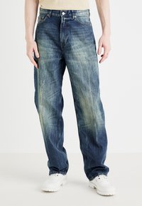 Vaqueros de denim azul con ajuste relajado, detalles desteñidos y acentos desgastados. Combinados con calzado blanco, sobre un fondo liso.