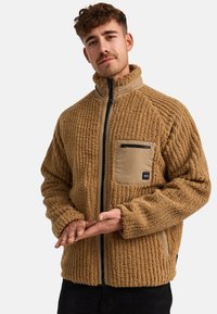 Chaqueta de forro polar marrón con un diseño texturizado y acanalado. Presenta un bolsillo con cremallera en color beige, cuello alto y detalles de cremallera en negro.