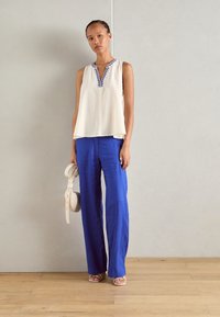 White sleeveless top with blue embroidery at the neckline, paired with loose blue linen pants. The model holds a cream-colored handbag.