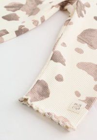Tissu côtelé crème avec imprimé vache marron clair, ourlet à bord ondulé et étiquette "milk club" dans un coin du textile.