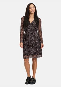 Langärmliges Wickelkleid aus transparentem schwarzem Stoff mit braunem Leopardenmuster, tailliertem Bund und geschichtetem Rock, kombiniert mit klobigen schwarzen Schuhen.