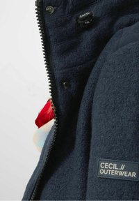 Navy blue fleece jacket with a zippered front, featuring a textured fabric and a logo patch. Includes a contrasting inner lining with patterns.