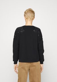 Sudadera negra con tejido texturizado, con cuello redondo y acentos con tachuelas en los hombros, combinada con pantalones beige.