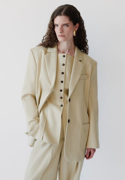 Γυναίκα με σγουρά μαλλιά φοράει μεγάλο μπεζ σακάκι, ταιριαστό μπλουζάκι με κουμπιά και παντελόνια, διακοσμημένα με αφηρημένα χρυσά σκουλαρίκια.