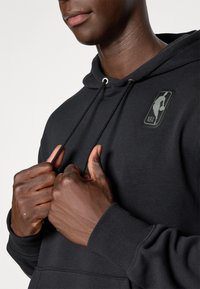 Personne ajustant les cordons d'un sweat à capuche noir NBA, montrant partiellement le visage et le torse avec le logo NBA sur la poitrine.