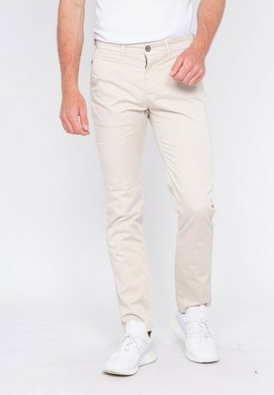 Homme portant un pantalon beige ajusté et des baskets blanches, debout devant un fond blanc uni.