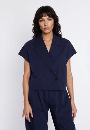 Kobieta o ciemnych, falowanych włosach, ubrana w granatową, krótką, dwurzędową marynarkę z krótkim rękawem i dopasowane spodnie, na białym tle.