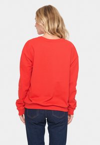 Felpa rossa con scollo tondo, taglio rilassato, maniche lunghe e polsini a costine, abbinata a jeans blu scuro. Texture morbida, senza motivi visibili.