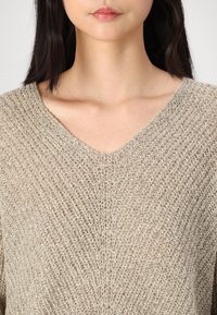 Femme portant un pull en tricot beige texturé avec un col en V, montrant les clavicules et une partie de ses cheveux foncés.