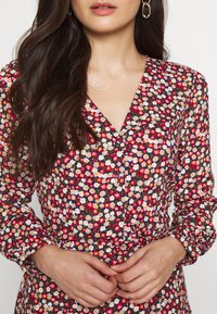 Abito floreale su sfondo blu navy con fiori vivaci rossi, rosa e bianchi. Presenta un collo a V, maniche lunghe e una vita cinte con un cinturino.