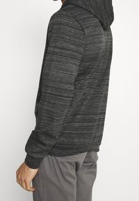 Hoodie grigio scuro con un design a righe testurizzato, dotato di cappuccio con cordino e polsini a costine. Indossato sopra pantaloni grigio chiaro.