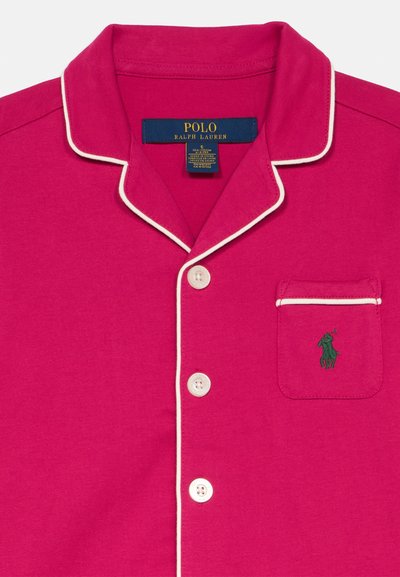 Pijama din bumbac roz cu guler tip notch, benzi albe și un logo verde pe buzunarul de la piept. Dispune de două butoane albe pe față.