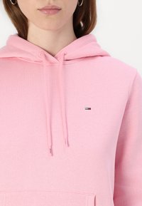 Hoodie rose clair en mélange de coton doux, avec une poche à l'avant et une capuche avec cordon de serrage. Petit logo brodé sur la poitrine dans des couleurs contrastées.