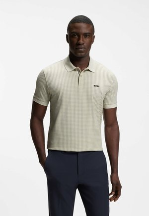 Ein Mann steht und trägt ein hellbeiges gemustertes Poloshirt mit „BOSS“-Logo und dunkelblaue Hose, Hände in den Taschen, vor einem schlichten weißen Hintergrund.