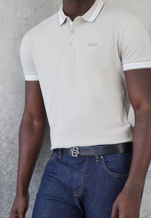 Mężczyzna ubrany w beżową koszulkę polo BOSS, włożoną do ciemnoniebieskich dżinsów, z czarnym paskiem z srebrną klamrą.