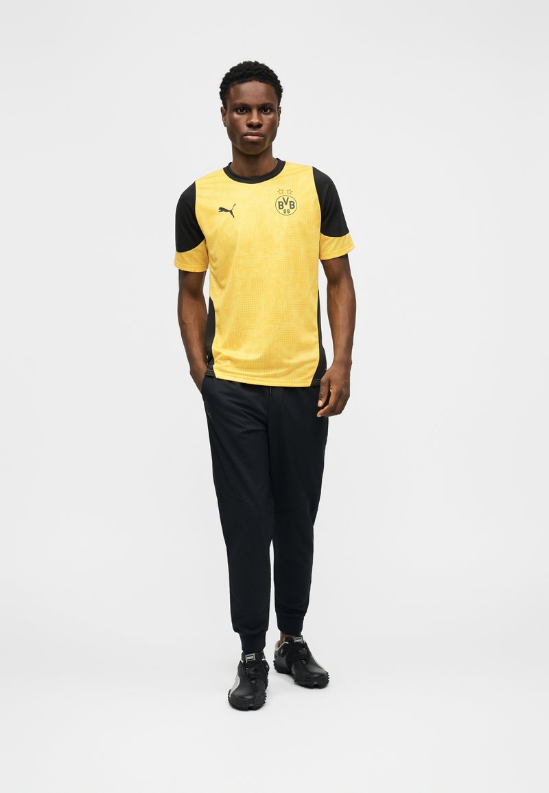 Muž stojící v žlutém a černém fotbalovém dresu Borussia Dortmund, černé kalhoty a černé sportovní boty na bílém pozadí.