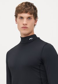 Jeune homme aux cheveux bruns courts portant un haut à col roulé noir ajusté à manches longues avec un petit logo blanc sur le col, sur fond blanc.