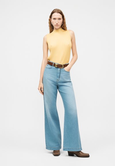 Femme portant un haut sans manches jaune côtelé, un jean large bleu clair avec une ceinture marron, et des chaussures à talons marron, debout avec une main dans la poche.