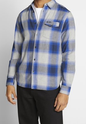 Homme portant une chemise à carreaux bleue, grise et blanche avec une poche poitrine et un pantalon noir, debout devant un fond uni.