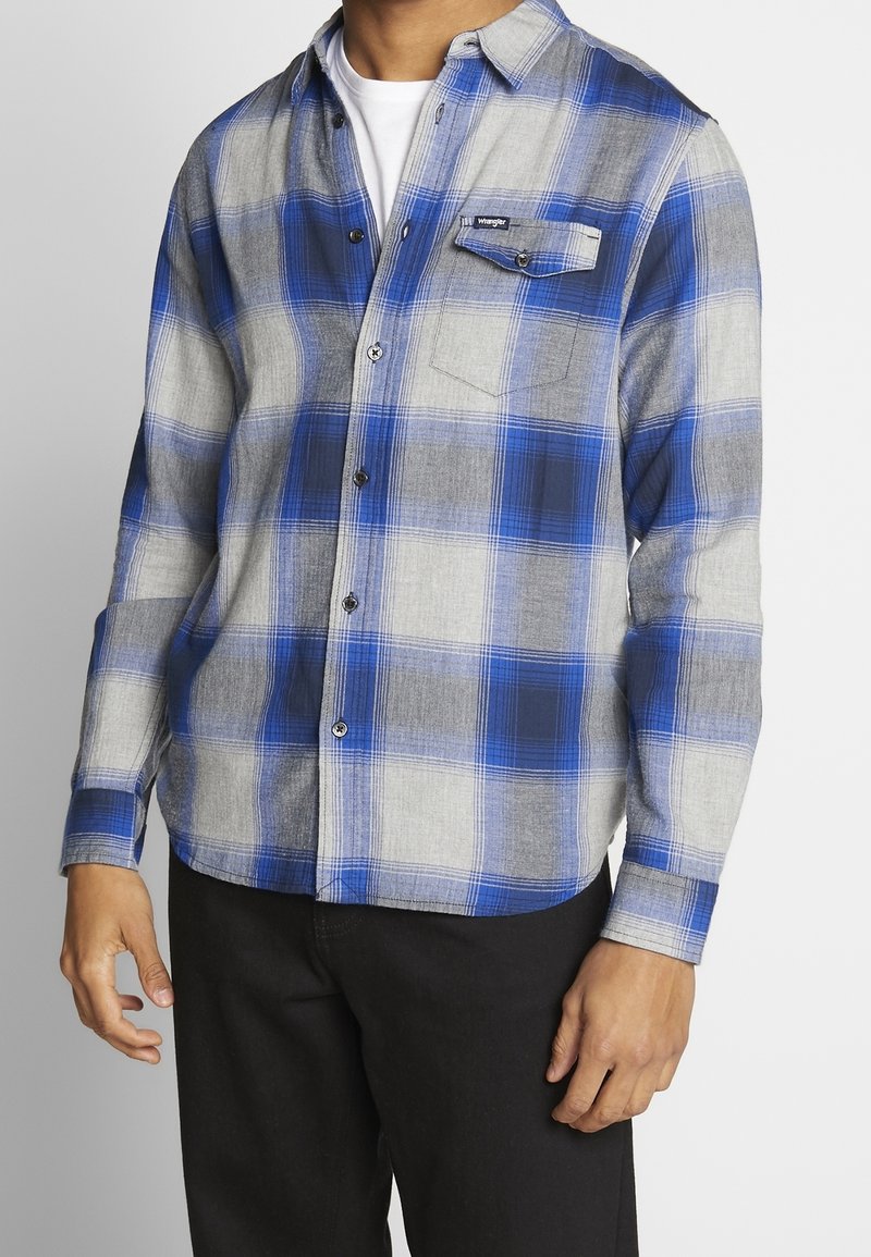 Homme portant une chemise à carreaux bleue, grise et blanche avec une poche poitrine et un pantalon noir, debout devant un fond uni.