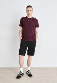 Burgundské tričko s malým logem, spárované s černými šortkami. Model má na sobě černé tenisky s bílými akcenty a bílé ponožky. Jednoduchý design, pohodlný střih.
