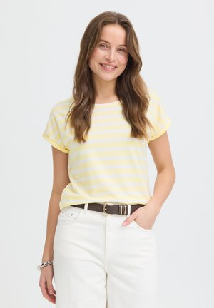 Žena s dlhými hnedými vlasmi, usmievajúca sa, oblečená v žlto-bielom pruhovanom tričku s krátkym rukávom, bielych nohaviciach a tmavom opasku, jedna ruka v vrecku.