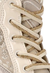 Primer plano de cordones metálicos en tonos beige y dorado pasados por ojales metálicos en los laterales texturizados de un zapato.