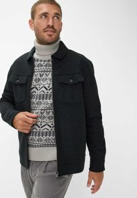 Schwarze Wollmischungjacke mit zwei vorderen Brusttaschen, getragen über einem cremefarbenen Rollkragenpullover mit einem blauen Wintermuster, kombiniert mit grauen Cordhosen.