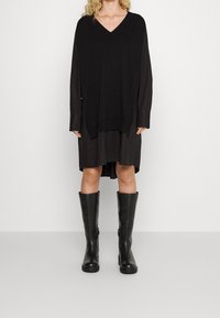 Schwarzes, übergroßes Strickkleid mit tiefem V-Ausschnitt, unter dem ein kürzeres Hemd getragen wird. Kombiniert mit kniefreien, eleganten schwarzen Gummistiefeln.