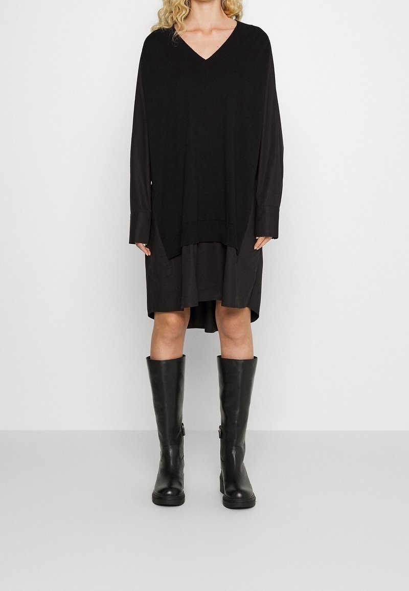 Schwarzes, übergroßes Strickkleid mit tiefem V-Ausschnitt, unter dem ein kürzeres Hemd getragen wird. Kombiniert mit kniefreien, eleganten schwarzen Gummistiefeln.