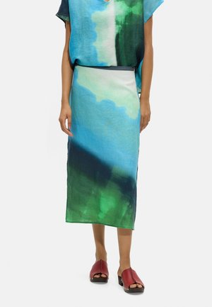 Kobieta ubrana w midi spódnicę z abstrakcyjnym wzorem w niebieskie, zielone i białe kolory, do tego dopasowany top oraz czerwone sandały na obcasie z odkrytymi palcami.