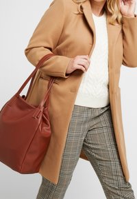 Bruine leren totebag met een gestructureerde afwerking, zacht vallend ontwerp, gecombineerd met een beige wollen jas en geruite broek.