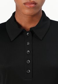 Crna polo majica s ravnim ovratnikom, šest dugmadi na prednjoj strani s crnim dugmadima i suptilnim logotipom na vratu. Glatka tekstura.