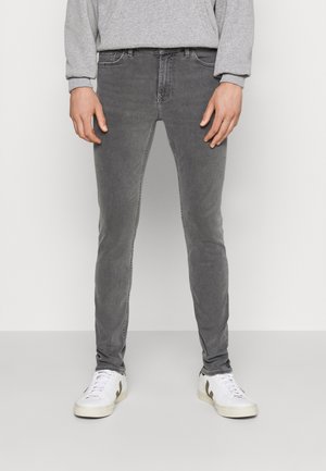 Person trägt eng anliegende graue Jeans, ein graues Langarmshirt und weiße Turnschuhe und steht vor einem einfarbigen Hintergrund.