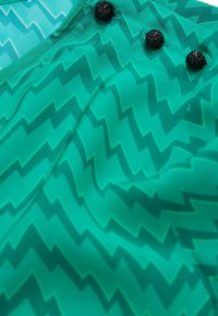 Green patterned textile featuring a zigzag design, with three black decorative buttons at the neckline, smooth texture, and lightweight material.