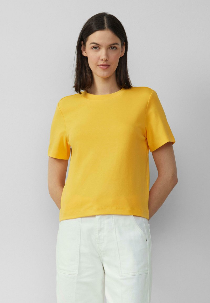 T-shirt jaune à manches courtes en coton avec un col rond, offrant une coupe décontractée et une texture douce. Associé à un pantalon blanc.
