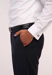 Weißes Hemd mit klassischem Kragen, langen Ärmeln und einem einzelnen Manschettenknopf mit lila Akzent. Getragen mit einem schwarzen Gürtel und marineblauen Hosen.