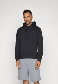 Svart hettegenser med kengurulomme, med en subtil okse-logo og Nike-merke, brukt sammen med grå treningsshorts.