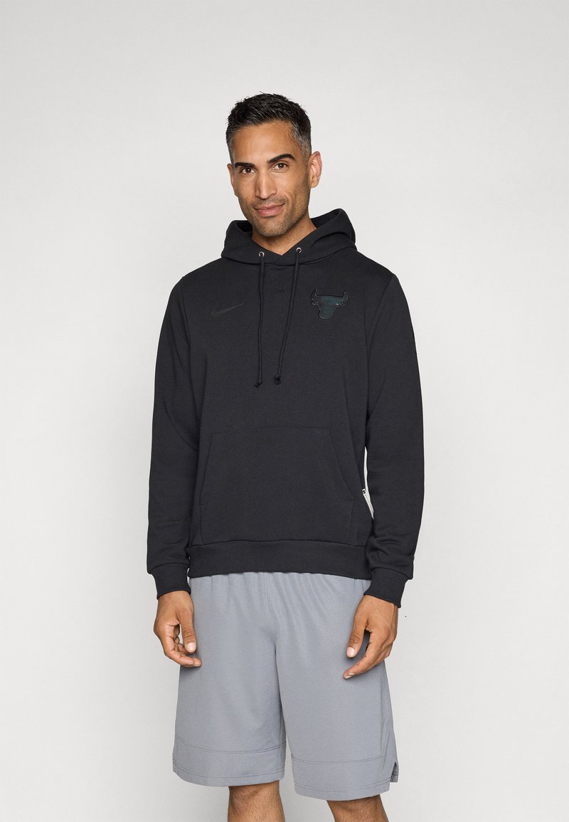 Felpa nera con cappuccio e tasca a marsupio, con un discreto logo di un toro e il marchio Nike, indossata con pantaloni sportivi grigi.