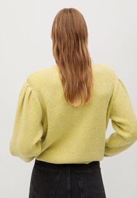 Gelbes strukturiertes Strickoberteil mit Puffärmeln, verziert mit kleinen glitzernden Details. Getragen mit schwarzen hochgeschnittenen Hosen.