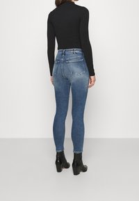 Mujer con jeans ajustados de color azul desgastado, camiseta de manga larga negra y botas negras hasta el tobillo, de pie de espaldas contra un fondo liso.