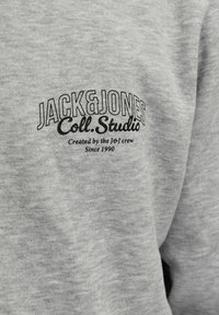 Grå stof med sort tekst "JACK & JONES Coll. Studio Created by the J&J crew Since 1990" trykt på brystområdet.