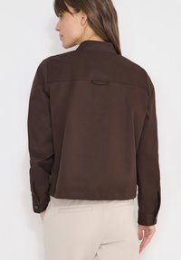 Femme aux longs cheveux bruns portant une veste marron foncé et un pantalon beige clair, vue de dos sur un fond blanc uni.