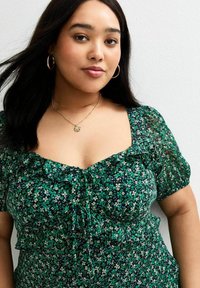 Groene bloempatron blouse met een vierkante halslijn, korte pofmouwen en ruches. Een gouden ketting en vergulde oorbellen complementeren de look.