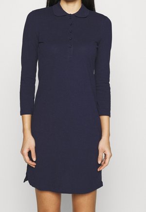 Femme portant une robe-polo bleu marine à manches longues avec des boutons sur le devant et un col arrondi, debout sur un fond uni.