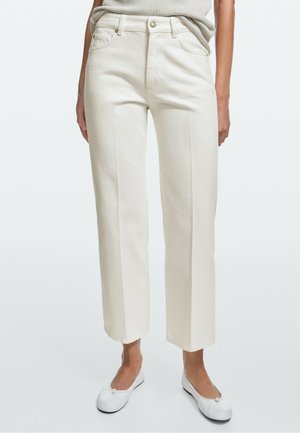 Donna che indossa pantaloni dritti color avorio, top grigio chiaro infilato e ballerine bianche, in piedi su uno sfondo chiaro uniforme.