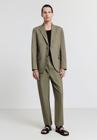 Uomo in piedi che indossa un abito verde oliva con camicia nera e sandali neri, su sfondo bianco uniforme.
