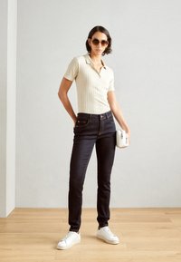 Beige ribbad polo skjorta med korta ärmar, mörka slim fit-jeans, vita sneakers och en liten vit väska. Modellen står på ett trägolv.